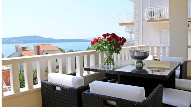 Un balcón con mobiliario de mimbre, una mesa con un jarrón de flores rojas y vistas al mar.