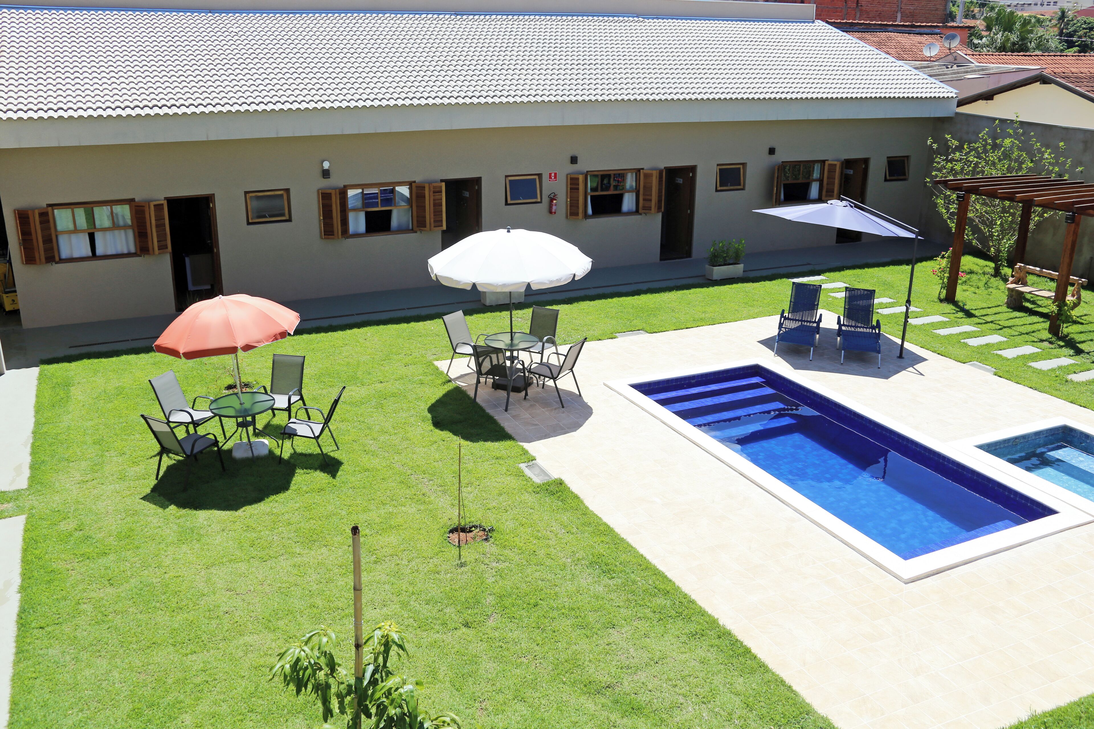 Zona de piscina con mesa y sillas, un espacio de estar cubierto y un edificio con varias ventanas y puertas.