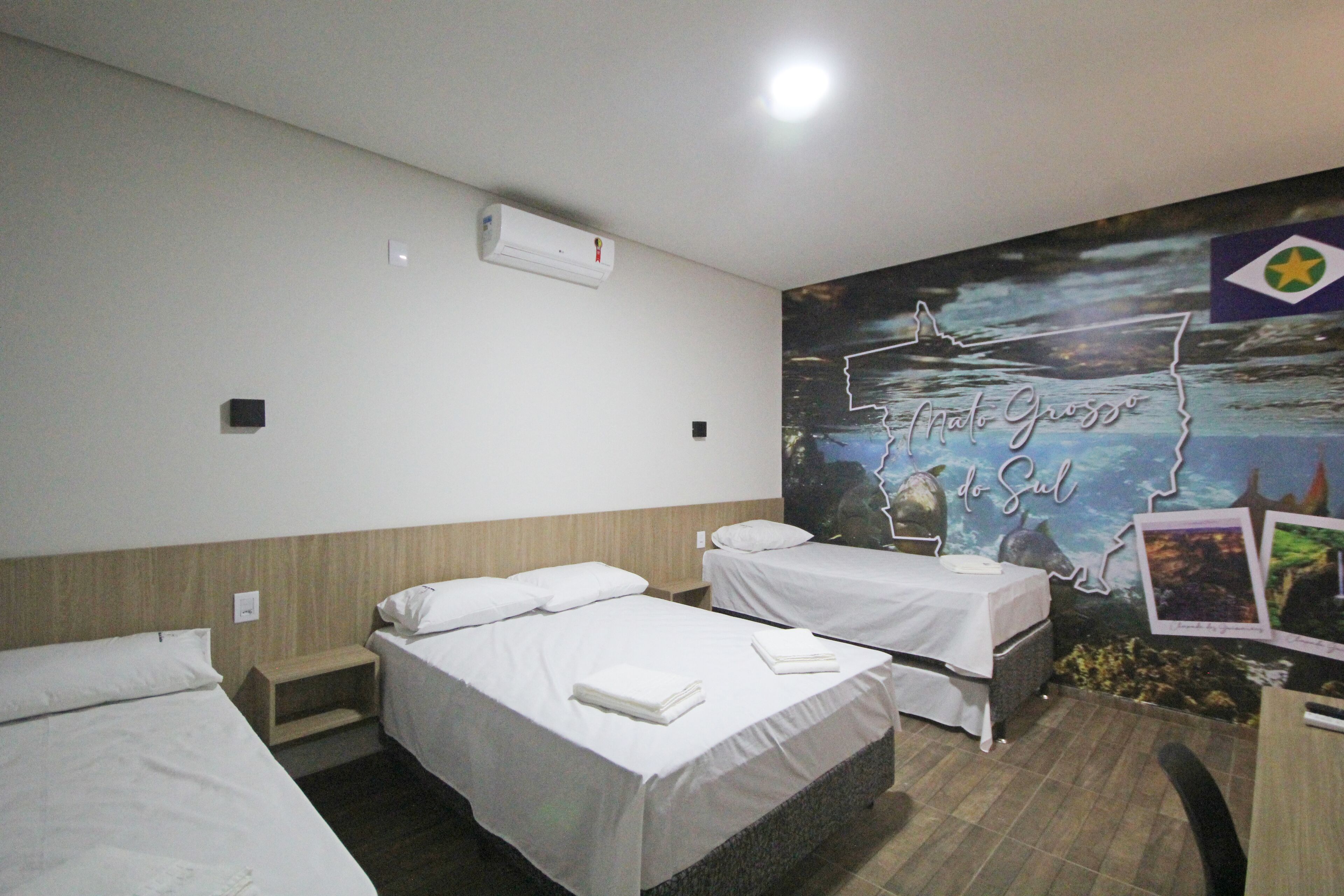 Une chambre d’hôtel avec deux lits, une tête de lit en bois, une fresque murale représentant un paysage côtier et un petit bureau avec une chaise.