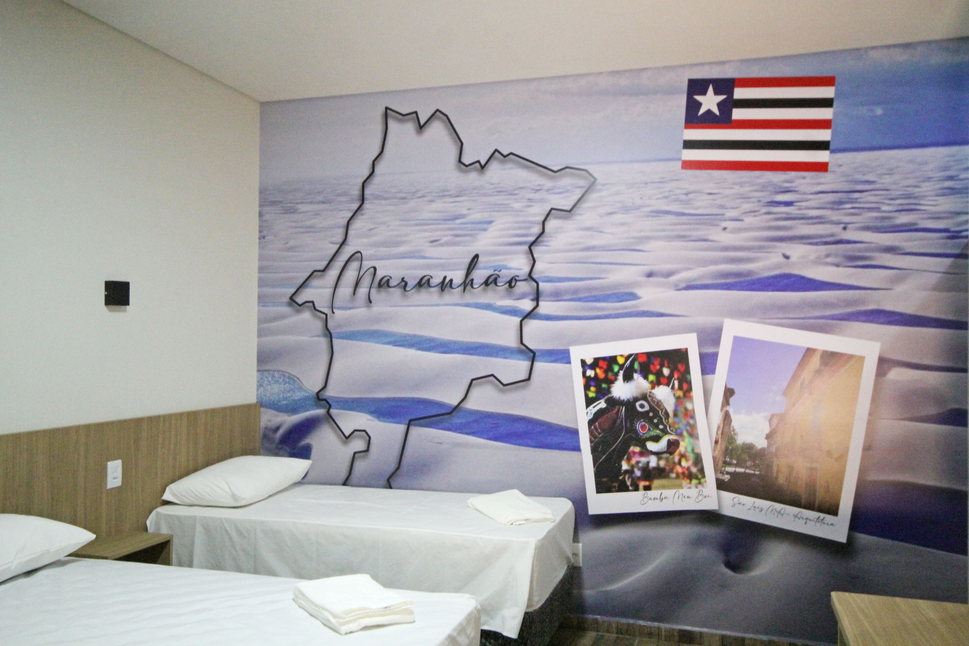 Una habitación de hotel con cama, mesita de noche y un mural en la pared que muestra un mapa con el nombre 'Maranhão' y una bandera.