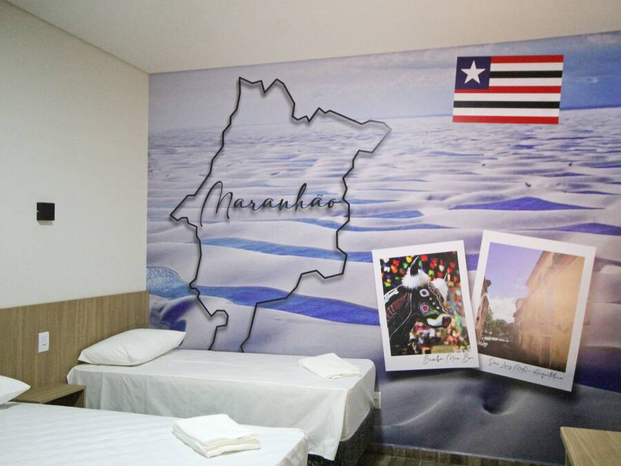 Una habitación de hotel con cama, mesita de noche y un mural en la pared que muestra un mapa con el nombre 'Maranhão' y una bandera.