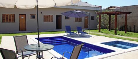 A swimming pool with a pergola and chairs, a covered patio area, and a building with doors and windows.