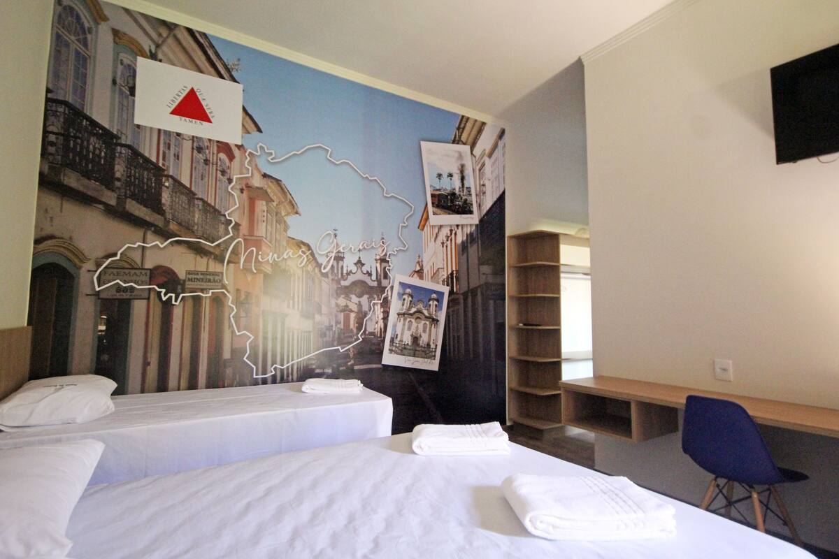 Ein Hotelzimmer mit zwei Betten, einem Schreibtisch, einem Stuhl und einer Wand mit einer Karte und Fotos.