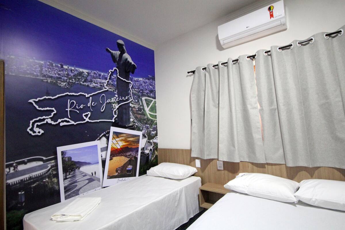 Ein Hotelzimmer mit zwei Betten, einem großen Wandgemälde von Rio de Janeiro und einem Fenster mit grauen Vorhängen.