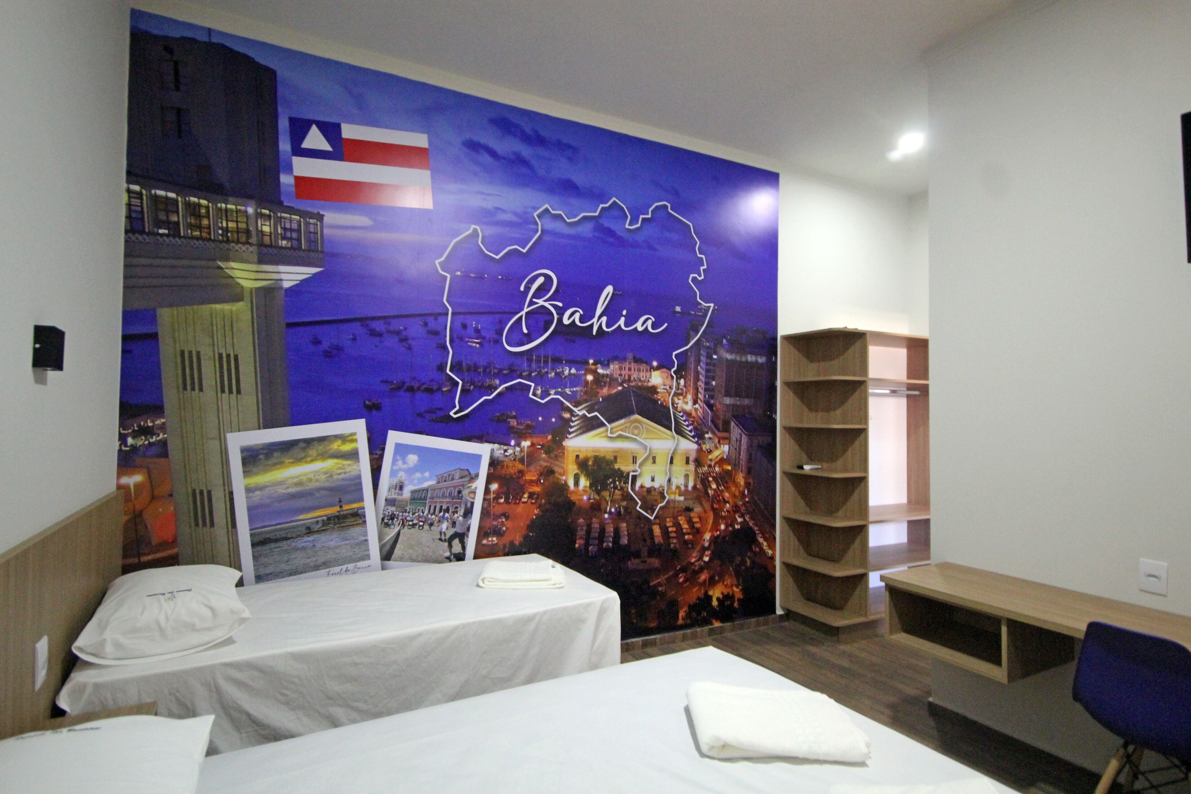 Camera d'albergo con due letti, un murale raffigurante un panorama urbano con la scritta 'Bahia', una bandiera e una libreria.