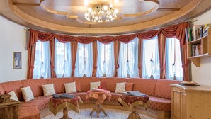 A cozy room with a curved wooden ceiling, a chandelier, a large window with curtains, a sofa with cushions, and a small table with a book. - Naturhotel Waldheim (Anterivo)