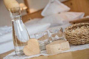 A clear glass bottle, two wooden heart-shaped objects, and two clear glasses on a wooden table with a woven basket. - Naturhotel Waldheim (Anterivo)