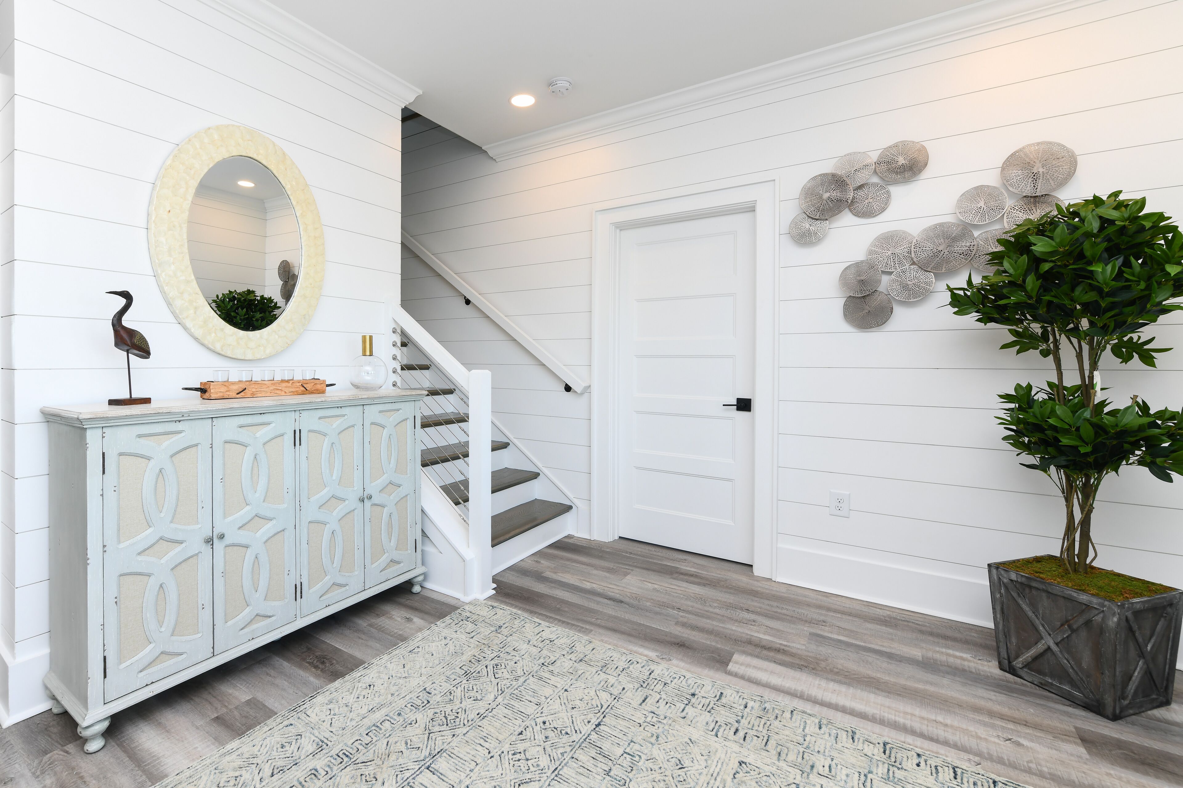 A hallway with a white wooden floor, a white staircase, a white door, a decorative mirror, a cabinet with a plant, and a potted plant.