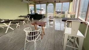 A spacious room with a high ceiling, multiple ceiling fans, and large windows offering a view of the outside. There are several tables and chairs arranged for dining or seating. - Tapasthali By Meera Hotels (Narendranagar)