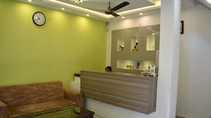A reception area with a sofa, a glass-top coffee table, a reception desk, and a ceiling fan. - Tapasthali By Meera Hotels (Narendranagar)