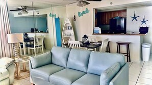 Una sala de estar con un sofá azul claro, una zona de comedor con una mesa y sillas, una cocina con refrigerador, y un ventilador de techo.