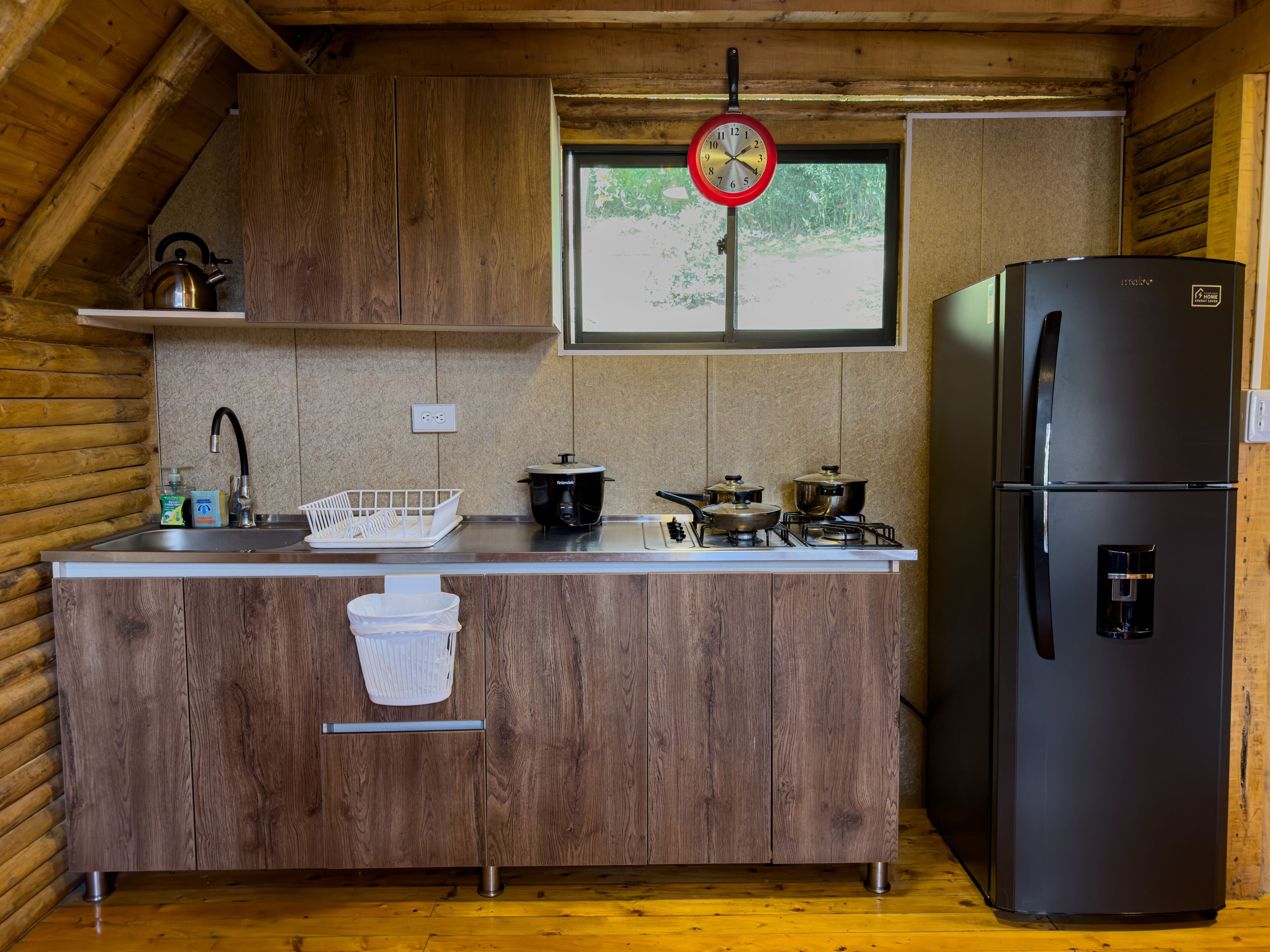 A compact kitchen with wooden cabinets, a stainless steel sink, a black refrigerator, and a window with a view of greenery.