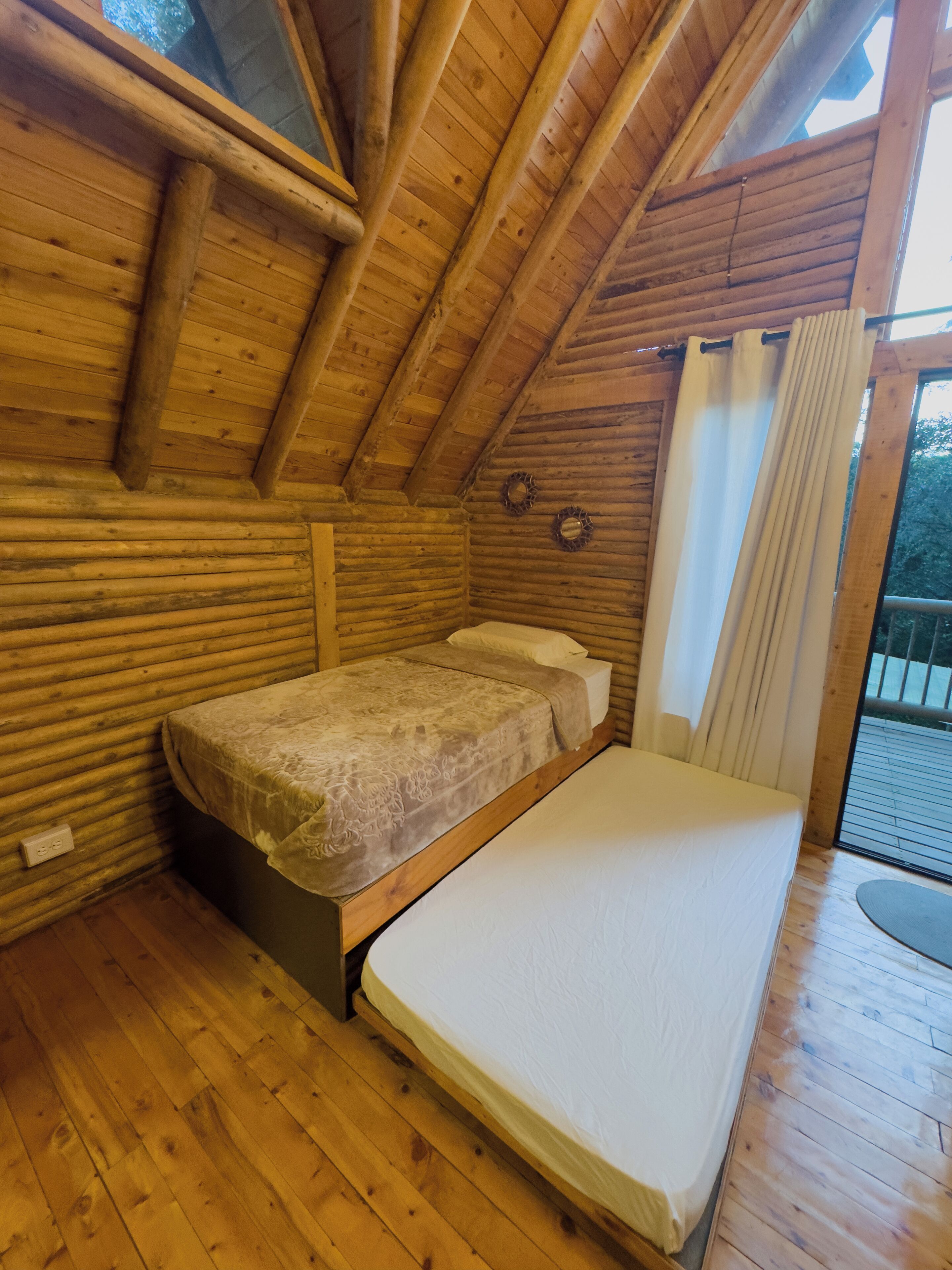 A wooden cabin room with a bed, a window with curtains, and a wooden floor.
