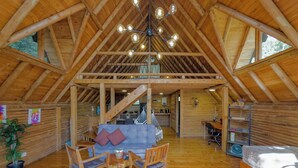 L’intérieur d’une cabane en rondins, avec un plafond en bois, un coin repas comprenant un canapé et des fauteuils, une cuisine à l’arrière-plan et un lit avec un matelas.