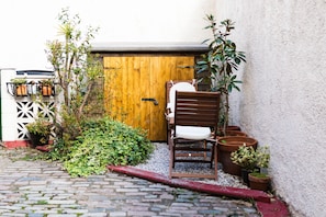 Un étroit sentier pavé de pierres menant à une porte en bois, bordé de plantes en pot et agrémenté d’un banc.