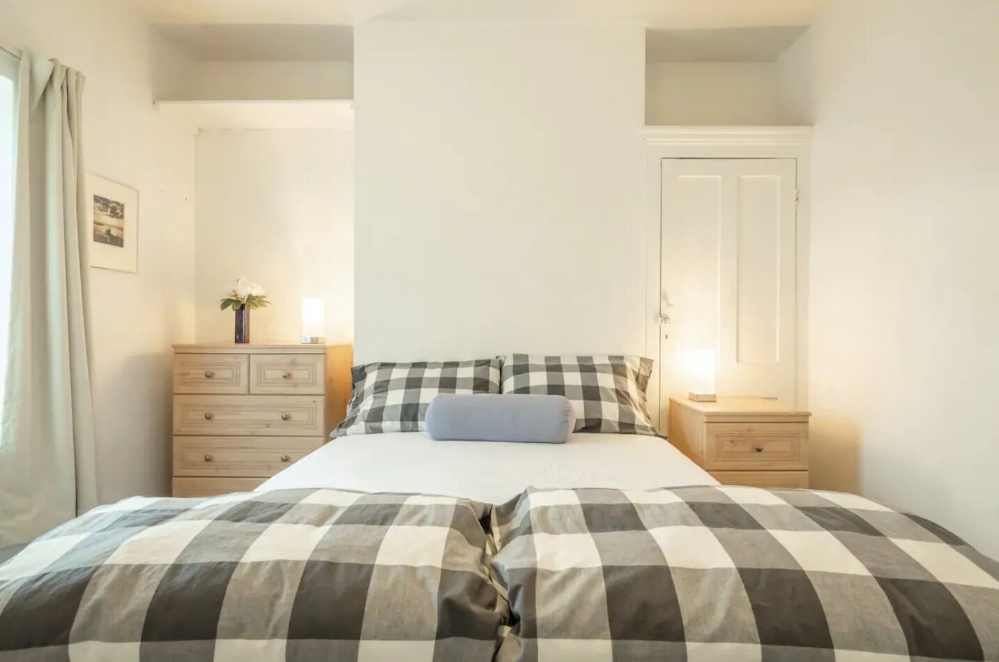 Een slaapkamer met een geruite sprei, een nachtkastje met lamp, een dressoir en een spiegel aan de muur.