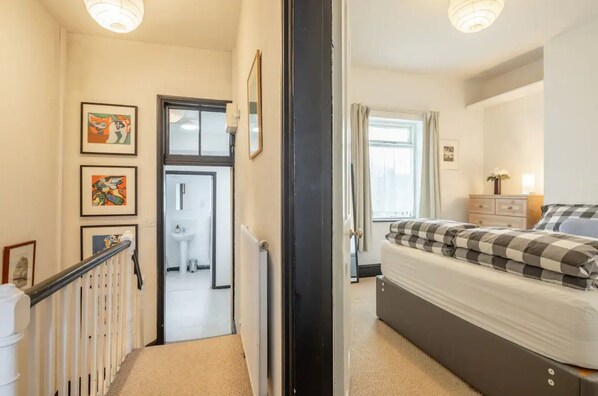 Ein Schlafzimmer mit Bett, Kommode und Fenster. Ein Flur führt zu einem Badezimmer mit Waschbecken und Toilette.