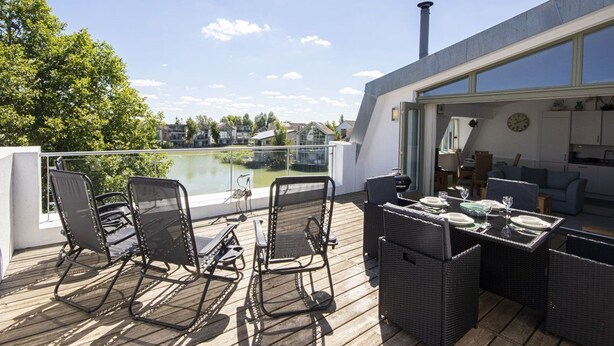 A modern outdoor deck with wicker furniture and a dining table, overlooking a lake and residential buildings. - Howells Mere 90, Crows Nest - Elevated Lake Views, Spa Access, Sleeps 4 (Somerford Keynes)