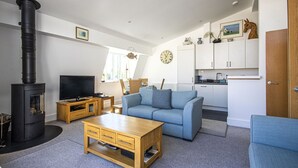 A modern living room with a black wood stove, a blue sofa, a wooden coffee table, and a TV on a wooden stand. - Howells Mere 90, Crows Nest - Elevated Lake Views, Spa Access, Sleeps 4 (Somerford Keynes)