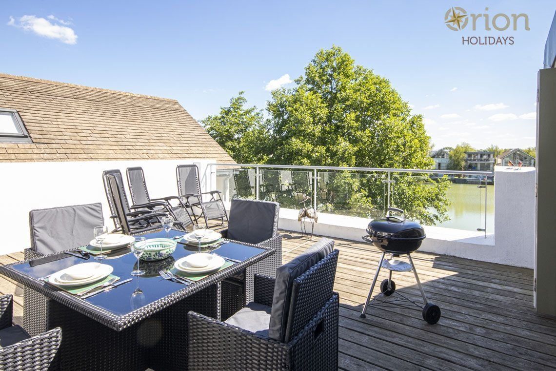 Ein Essbereich im Freien mit einem Tisch für vier Personen, umgeben von Korbstühlen, einem Grill und Blick auf einen See.