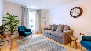 Un salón moderno con un sofá gris, un sillón azul, un reloj de pared redondo y una planta grande en maceta.