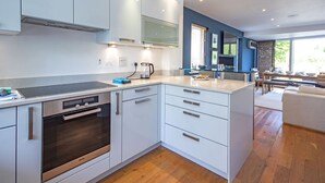 A modern kitchen with white cabinetry, a built-in oven, and a dining area with a wooden table and chairs. - Clearwater 67, Ortolan Villa (P) - Lakeside House, Hot Tub, Spa Access, Sleeps 8 (Somerford Keynes)
