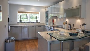 A modern kitchen with white cabinetry, a glass island counter, and a view of greenery outside. - Clearwater 67, Ortolan Villa (P) - Lakeside House, Hot Tub, Spa Access, Sleeps 8 (Somerford Keynes)