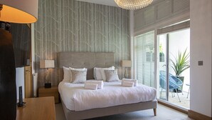 A modern bedroom with a large bed, two bedside tables, a chandelier, and a view of the outdoors through large sliding doors. - Clearwater 67, Ortolan Villa (P) - Lakeside House, Hot Tub, Spa Access, Sleeps 8 (Somerford Keynes)