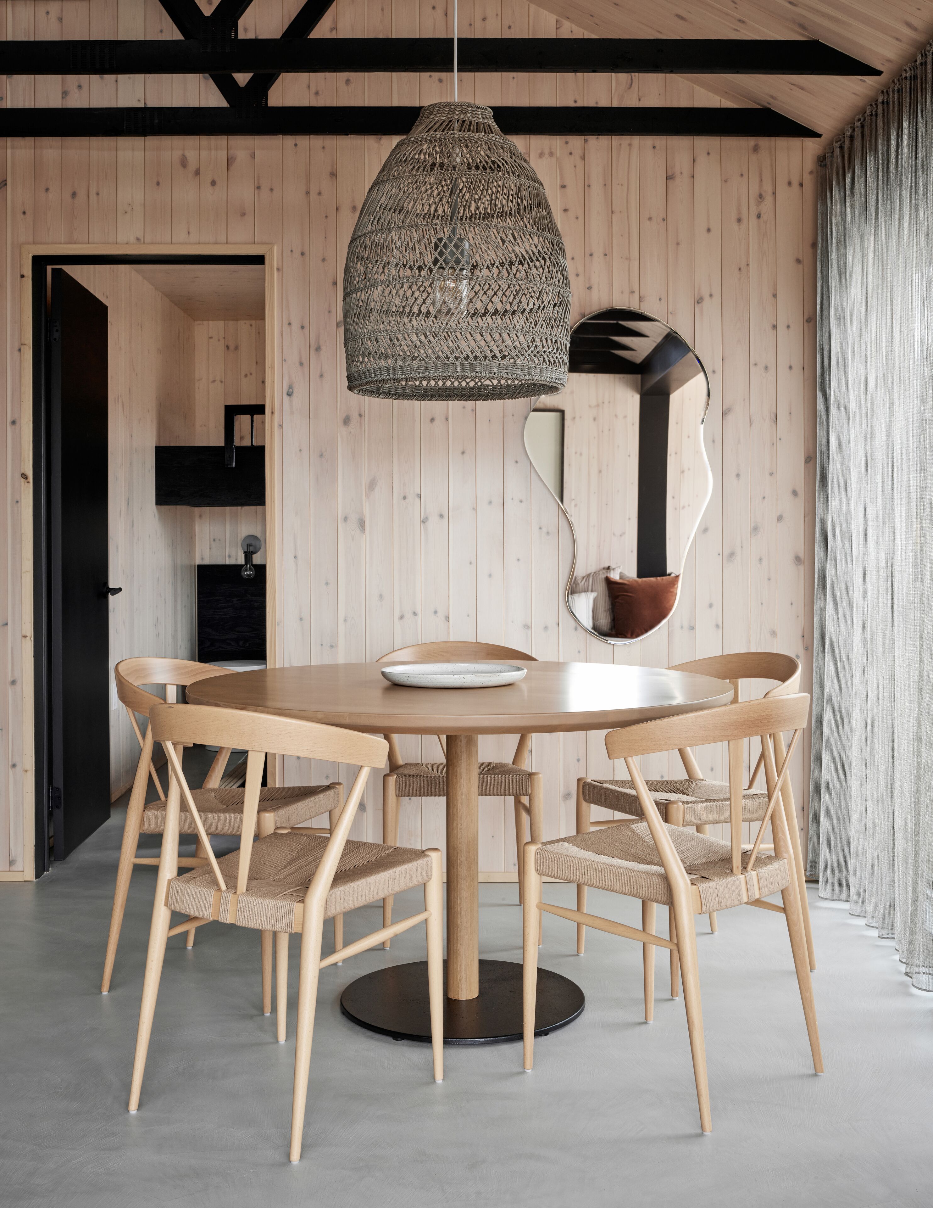 Un coin repas comprenant une table ronde en bois, six chaises, un luminaire suspendu en fibres tressées et un miroir moderne.