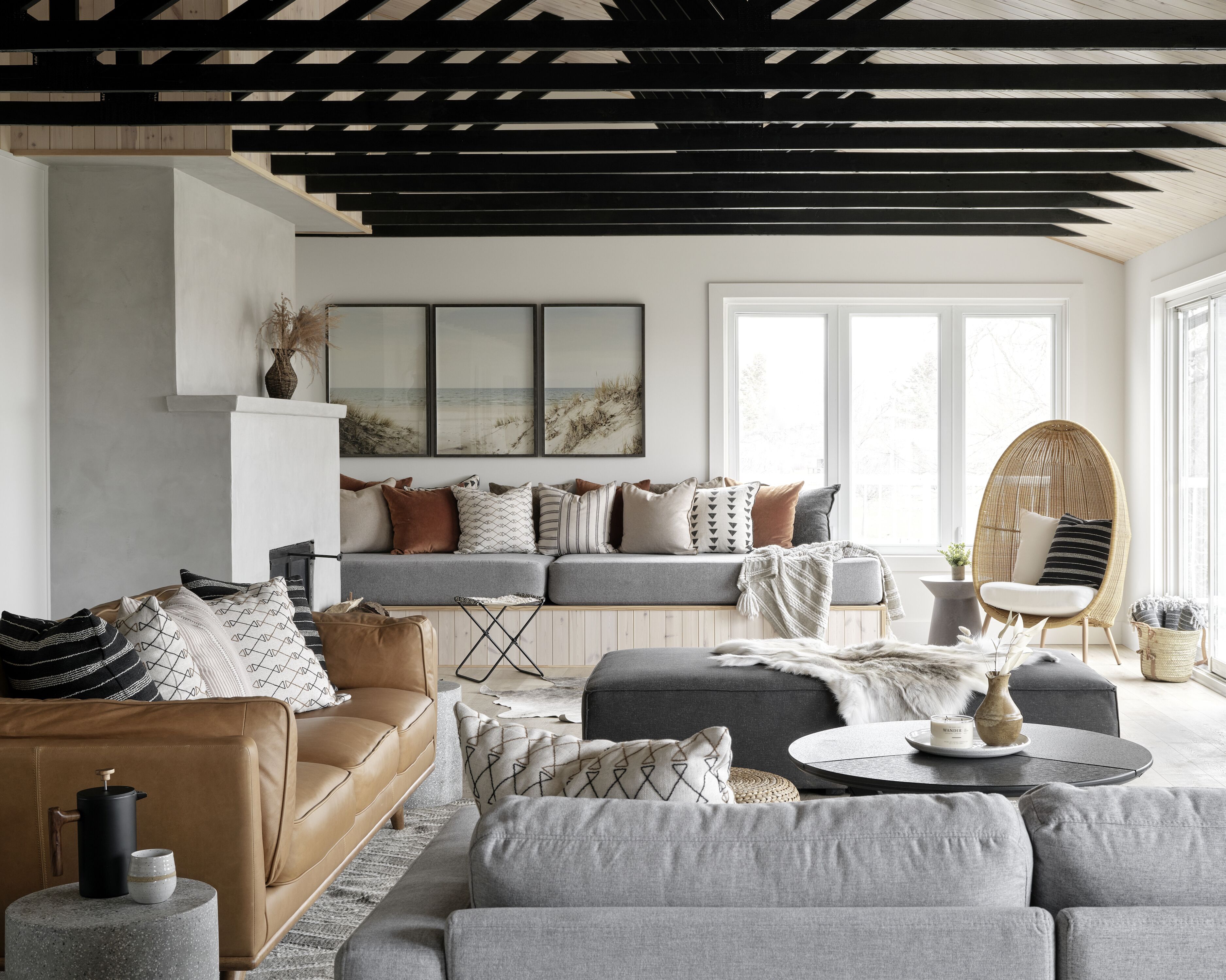 Een moderne woonkamer met een bank, fauteuils en een salontafel. Er is een open haard, een groot raam en een plafond met zichtbare balken.