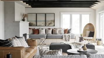 Een moderne woonkamer met een bank, fauteuils en een salontafel. Er is een open haard, een groot raam en een plafond met zichtbare balken.