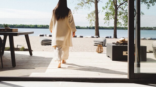 A person walking towards a beach from a room with a large window overlooking the water. - Wander the Resort (Prince Edward)
