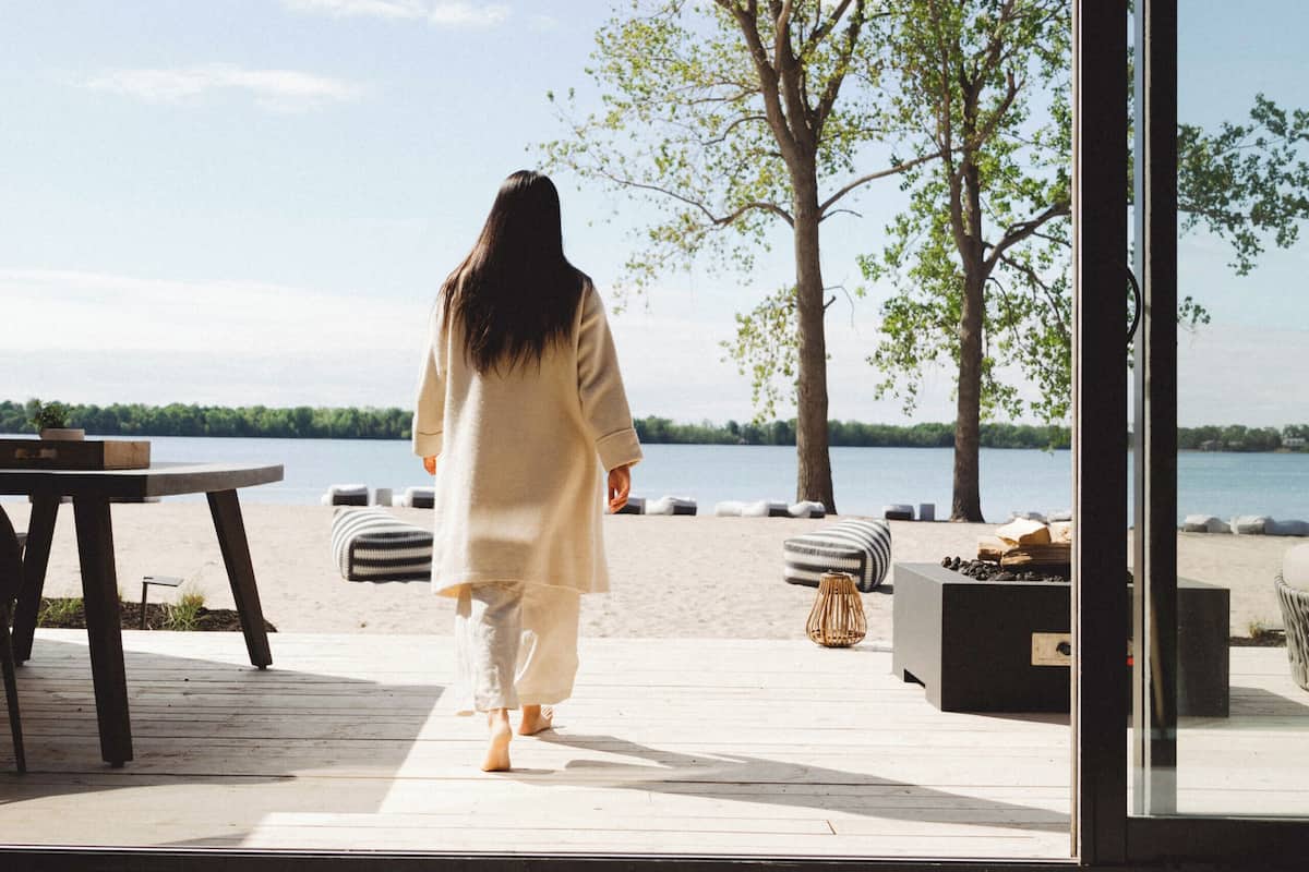 A person walking towards a beach from a room with a large window overlooking the water.