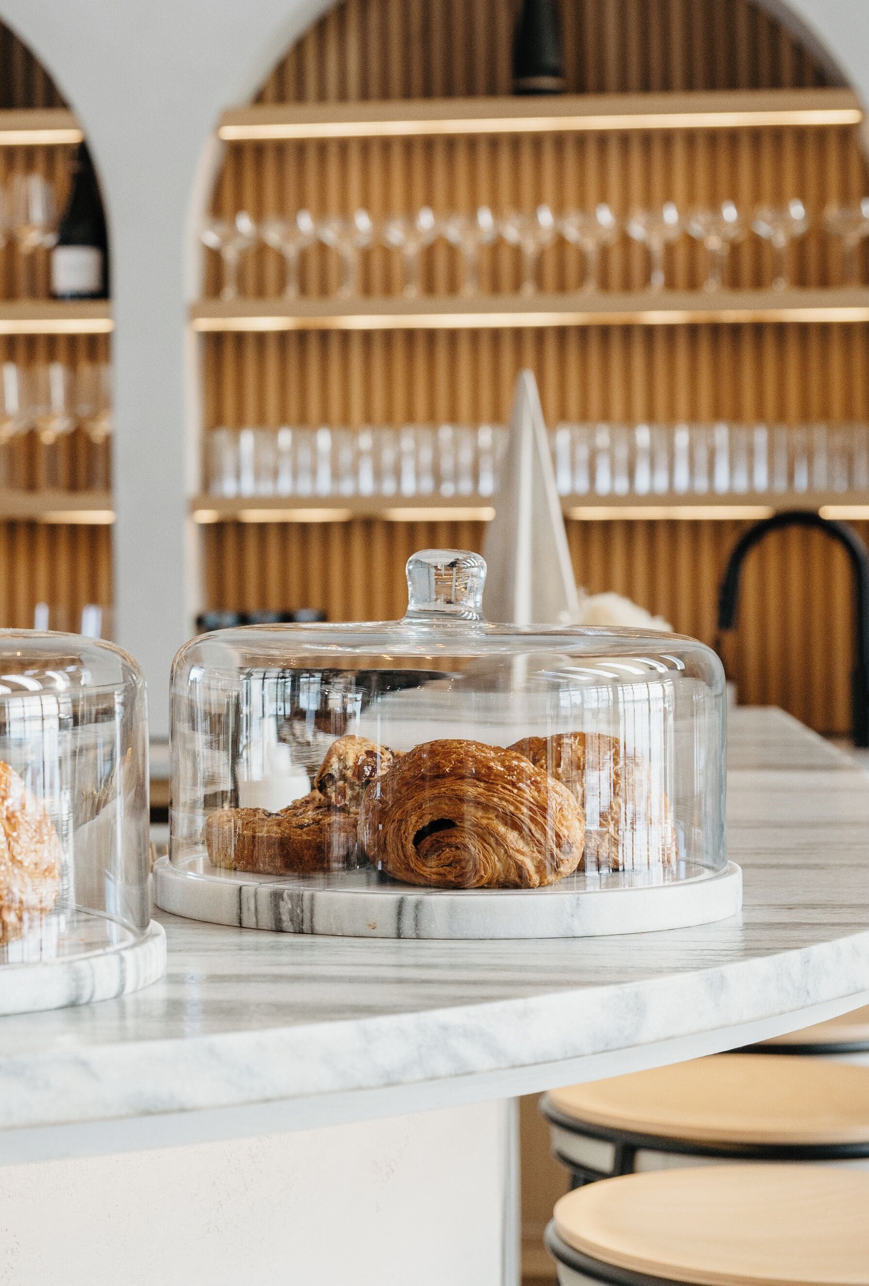 Une vitrine de boulangerie présentant des pâtisseries sous des cloches de verre, avec des comptoirs en marbre et des étagères en bois en arrière-plan.
