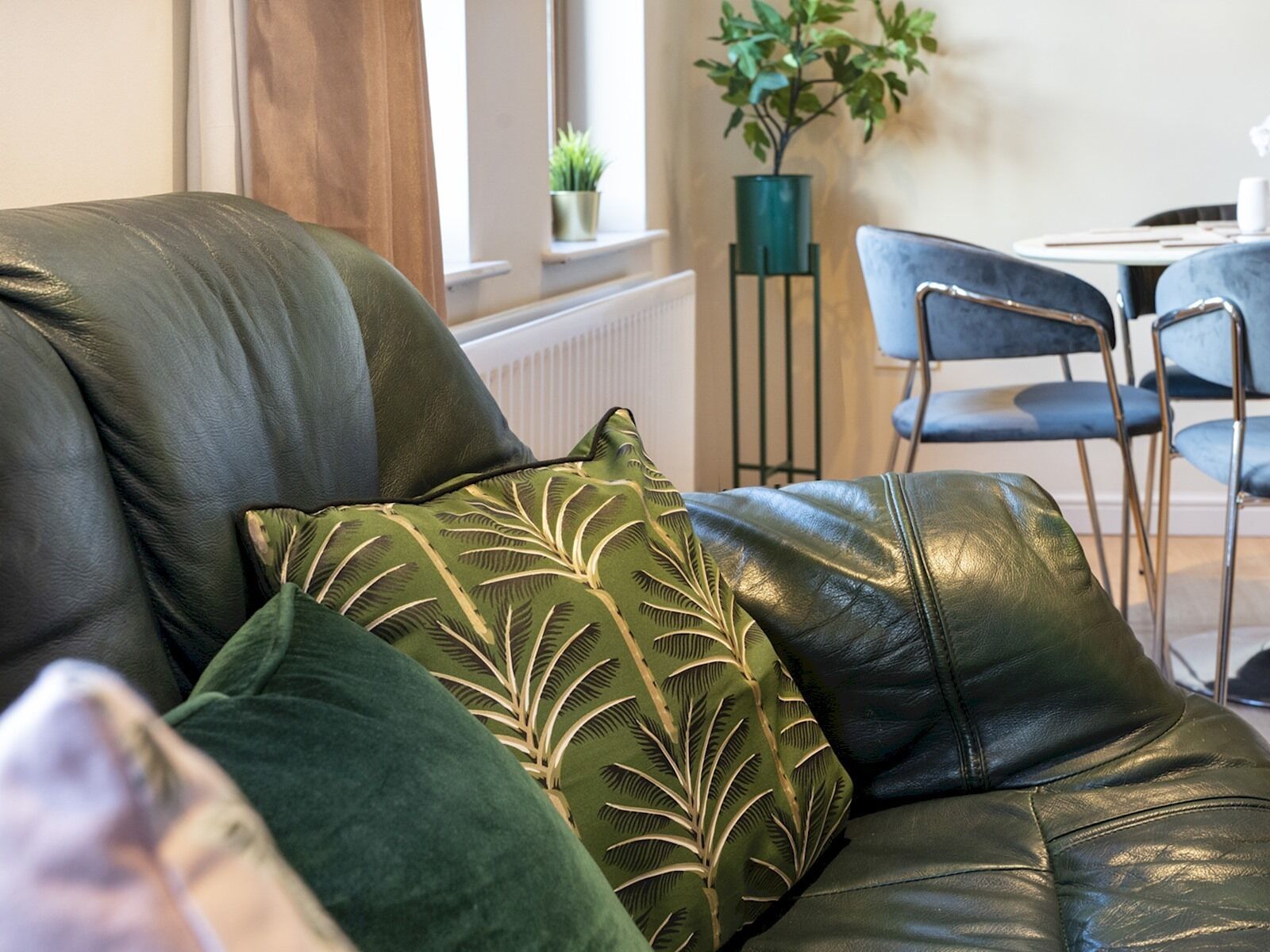 A leather sofa with green cushions in a room with a dining area and a potted plant.