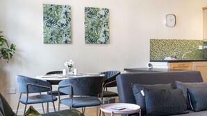 A modern living room with a grey sofa, blue chairs, and a small round table. There are two wall-mounted paintings with a tropical leaf pattern. A kitchen area with a green tiled backsplash and a clock is visible in the background. - Stunning 2BR Apt with Balcony Next to Queens Uni - Pass the Keys (Belfast)