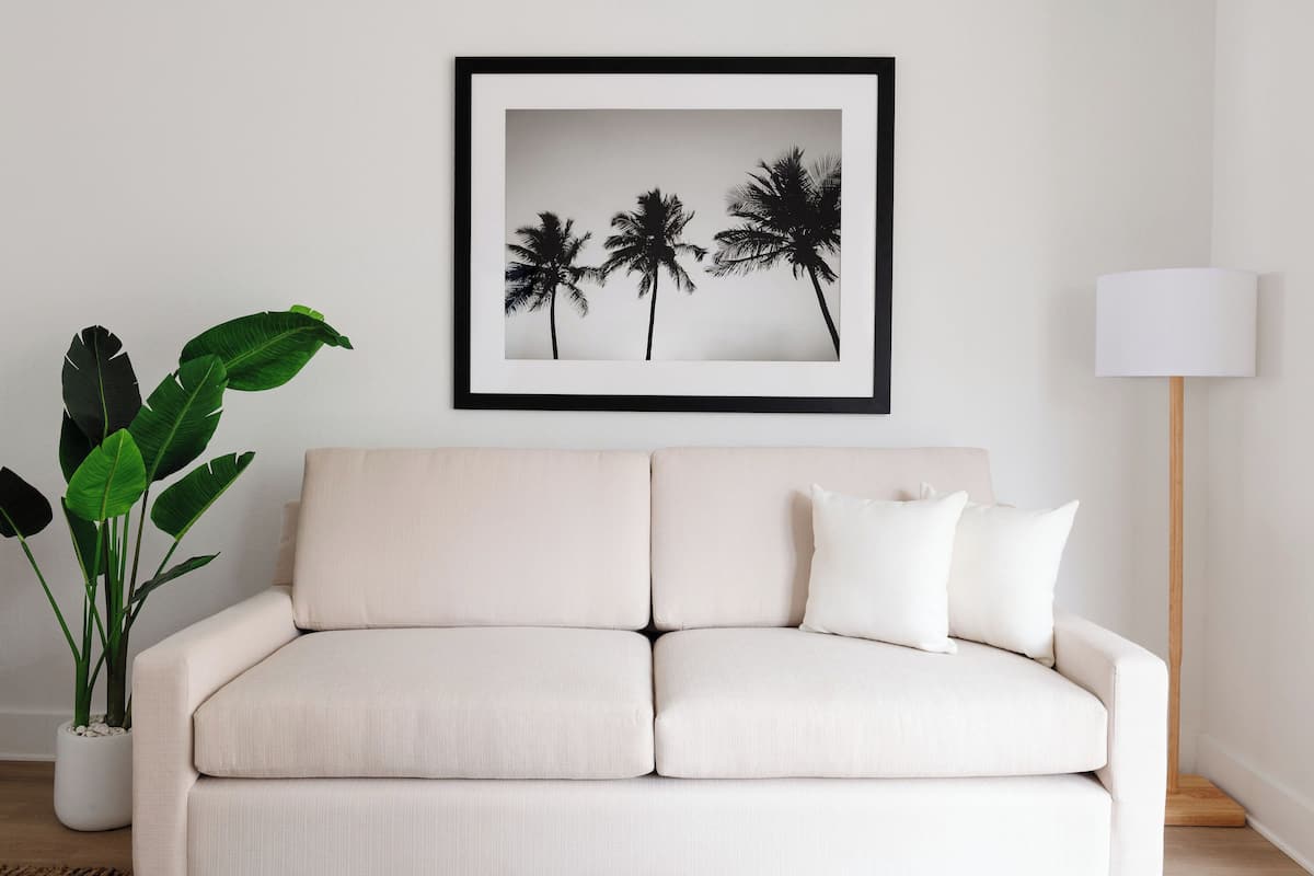 A room with a beige sofa, a framed picture of palm trees, a floor lamp, and a potted plant.