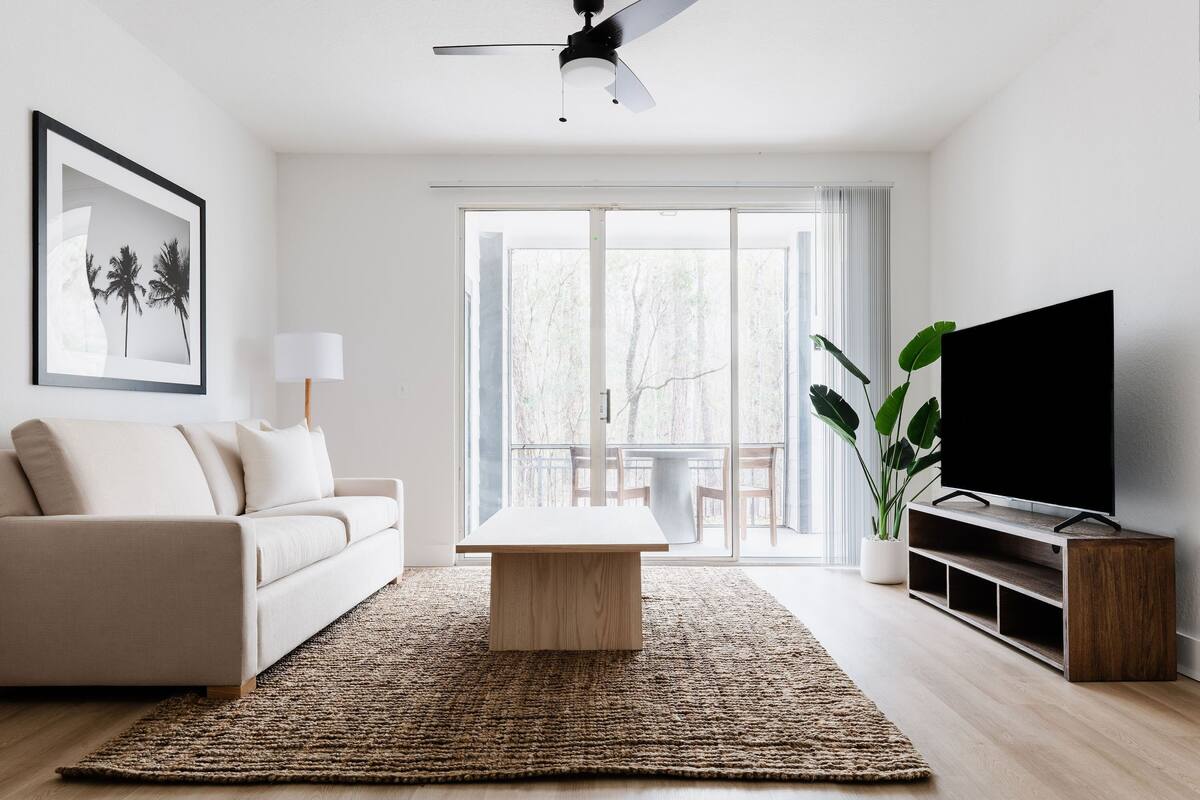 A modern living room with a sofa, a coffee table, a TV on a wooden stand, and a large window with a view of trees.