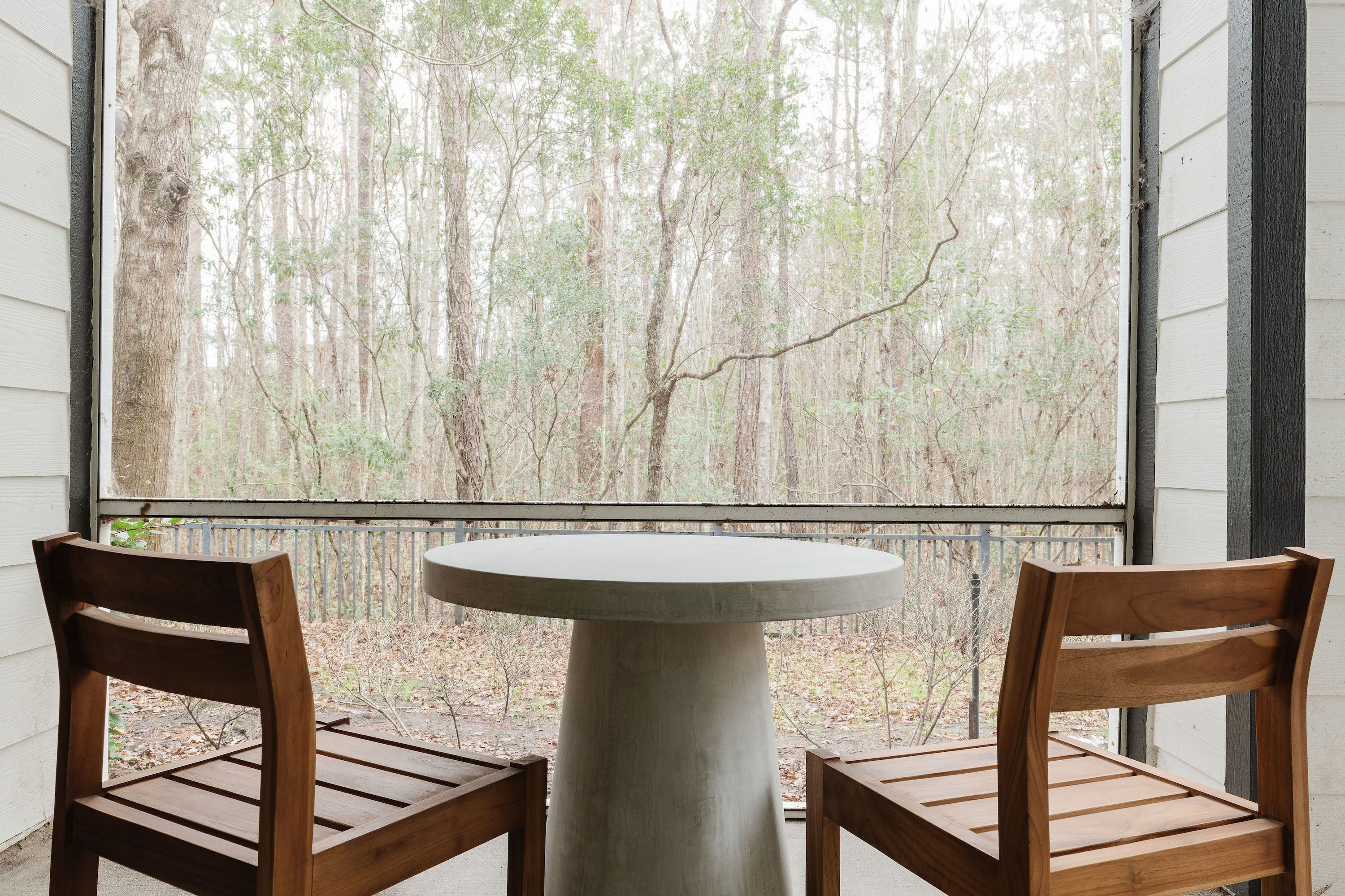 Une table ronde, accompagnée de deux chaises en bois, se trouve dans une pièce dotée d’une grande fenêtre donnant sur une forêt.
