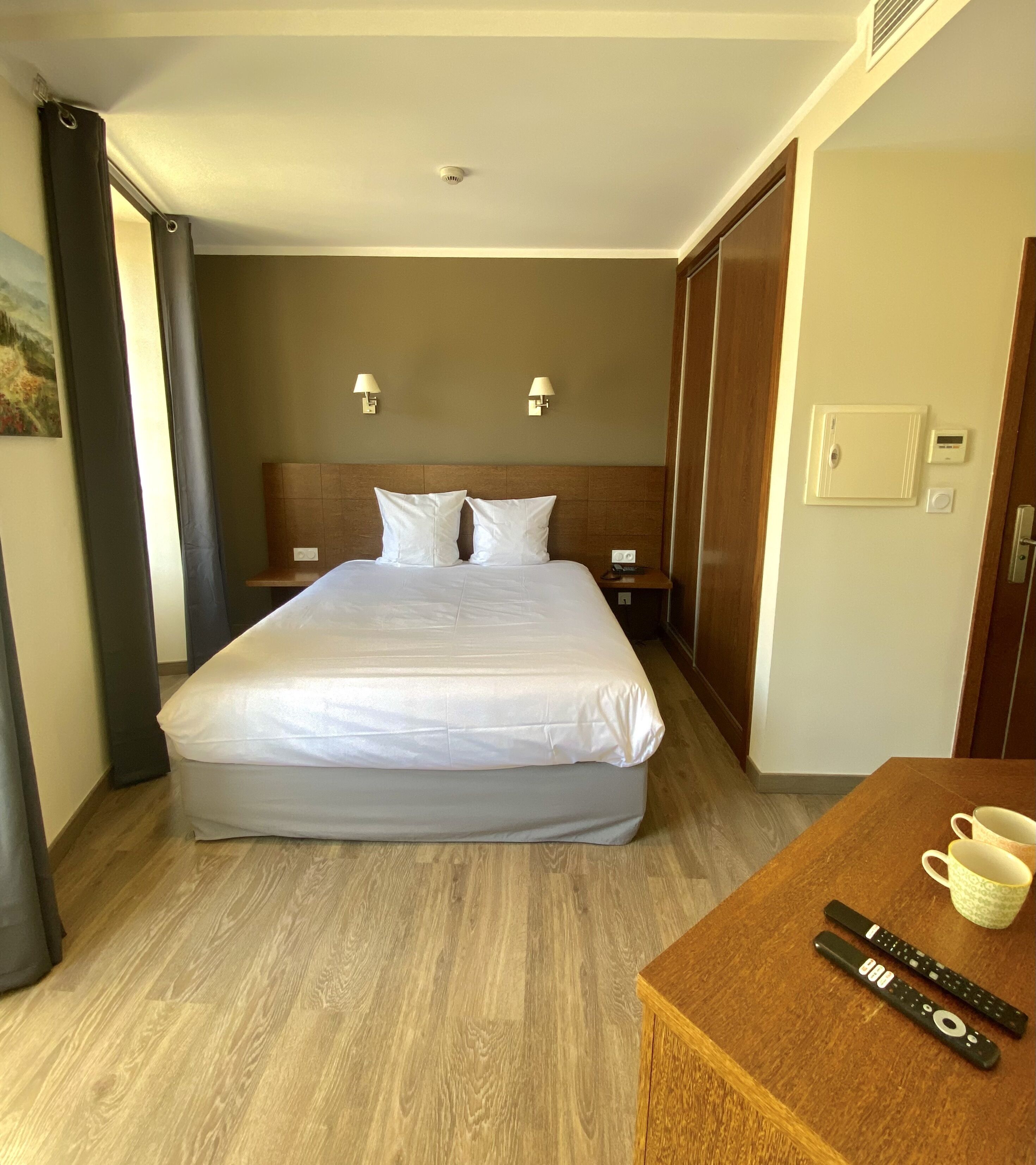 A hotel room with a large bed, two bedside tables, a wooden desk with a cup and remote controls, and a painting on the wall.