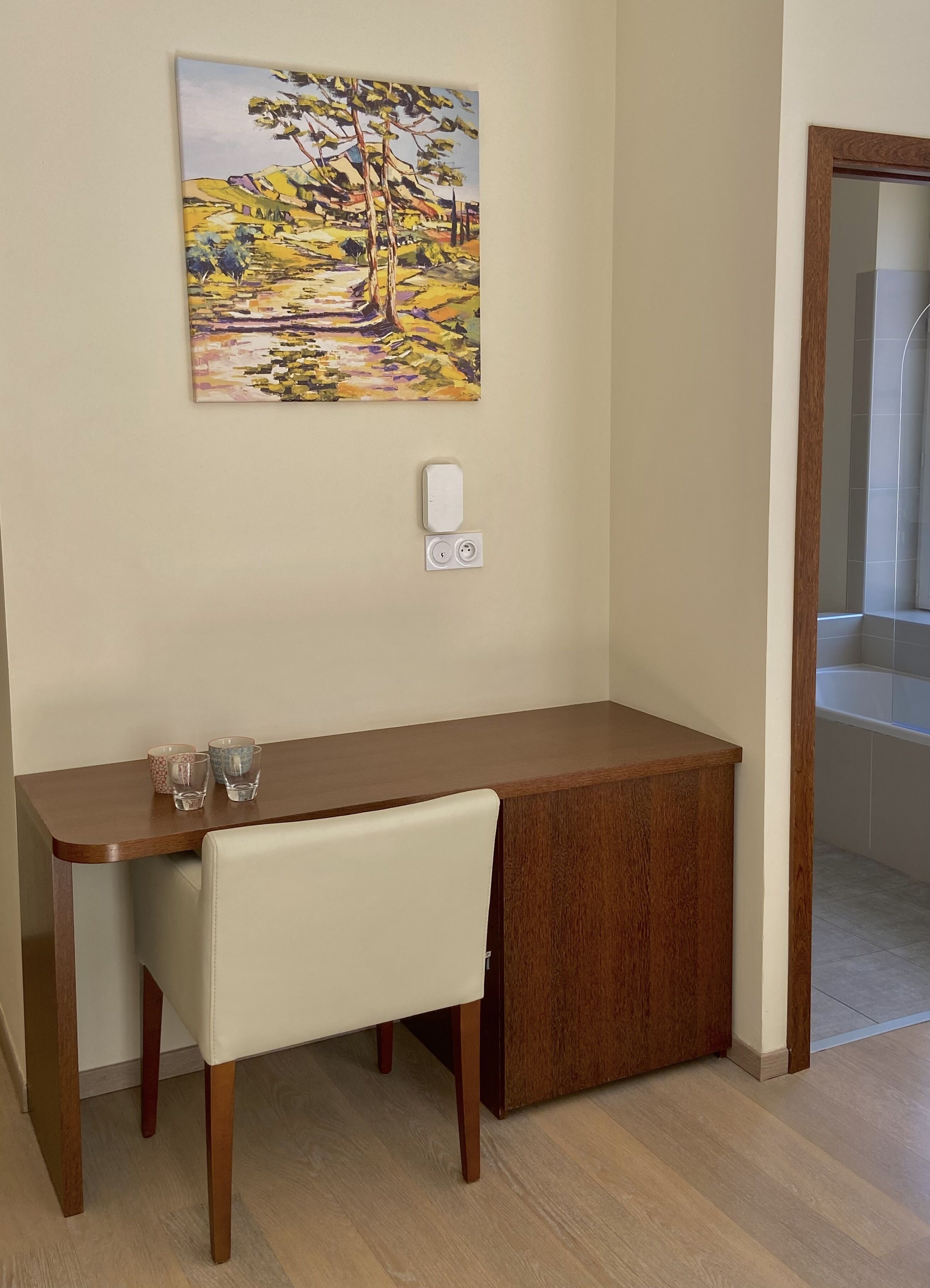 A small, modern room with a wooden desk, a chair, and a painting on the wall. There is a power outlet and a light switch on the wall.