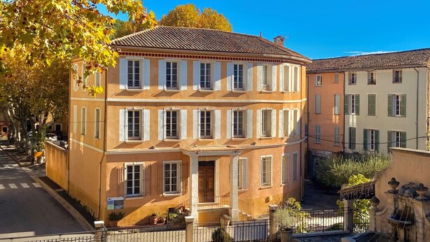 A multi-story building with a traditional architectural style, featuring a central entrance with a staircase, a courtyard with a fountain, and a street view with trees and parked cars. - Hôtel La Falaise (Cotignac)