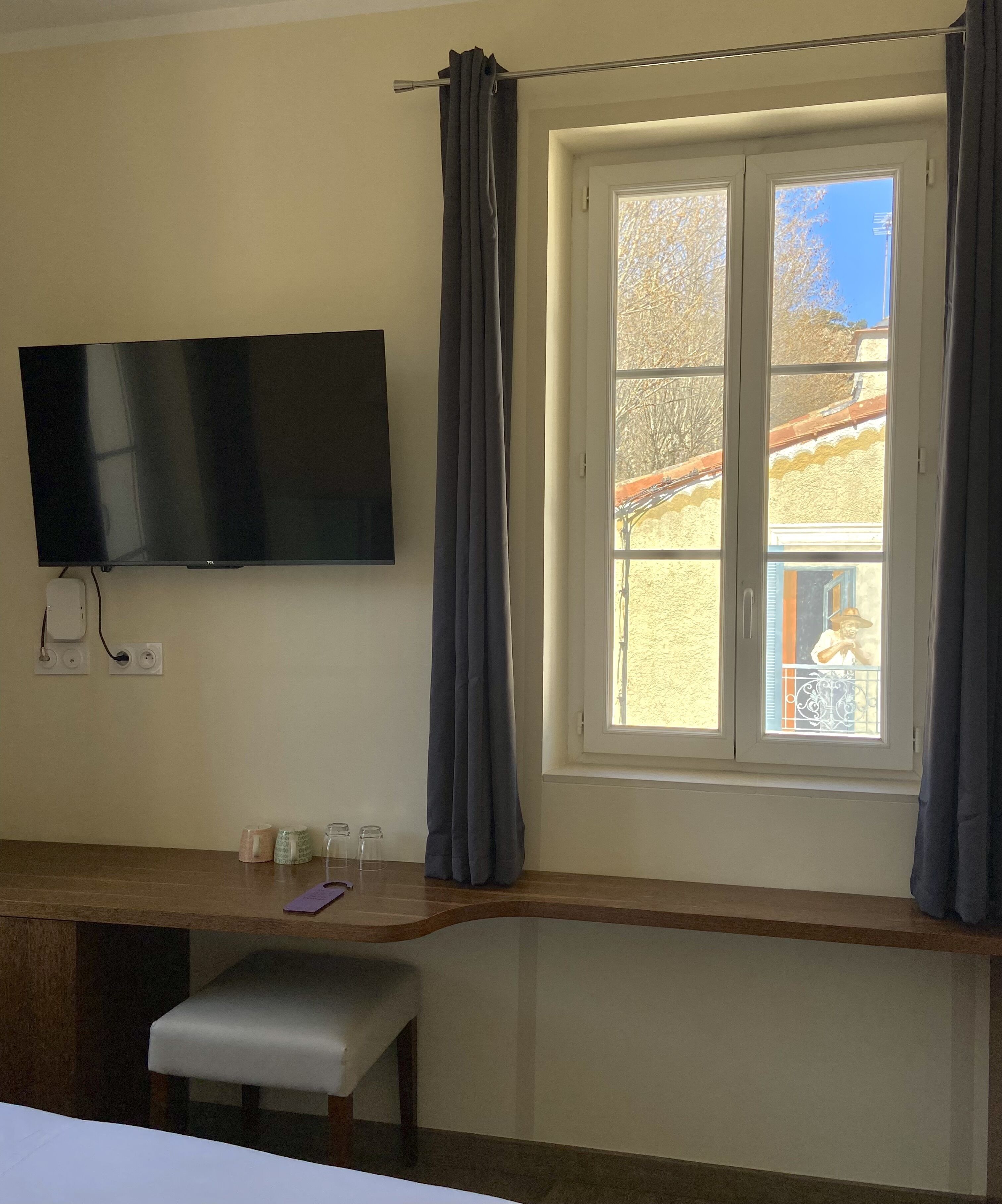 Una habitación con una televisión montada en la pared, una ventana con cortinas, un banco y una pequeña mesa con objetos encima.