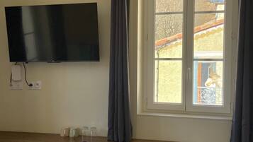 Una habitación con una televisión montada en la pared, una ventana con cortinas, un banco y una pequeña mesa con objetos encima.