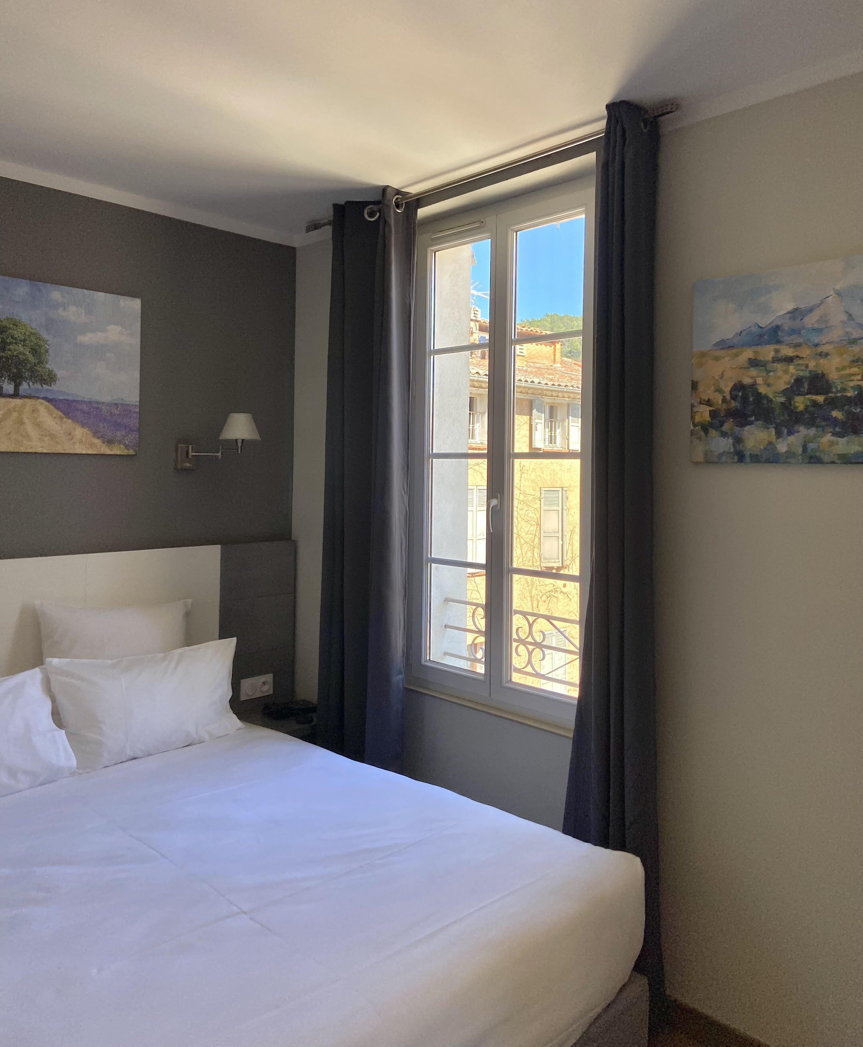 A neatly made bed with white linens, a wall-mounted lamp, and a window with curtains.