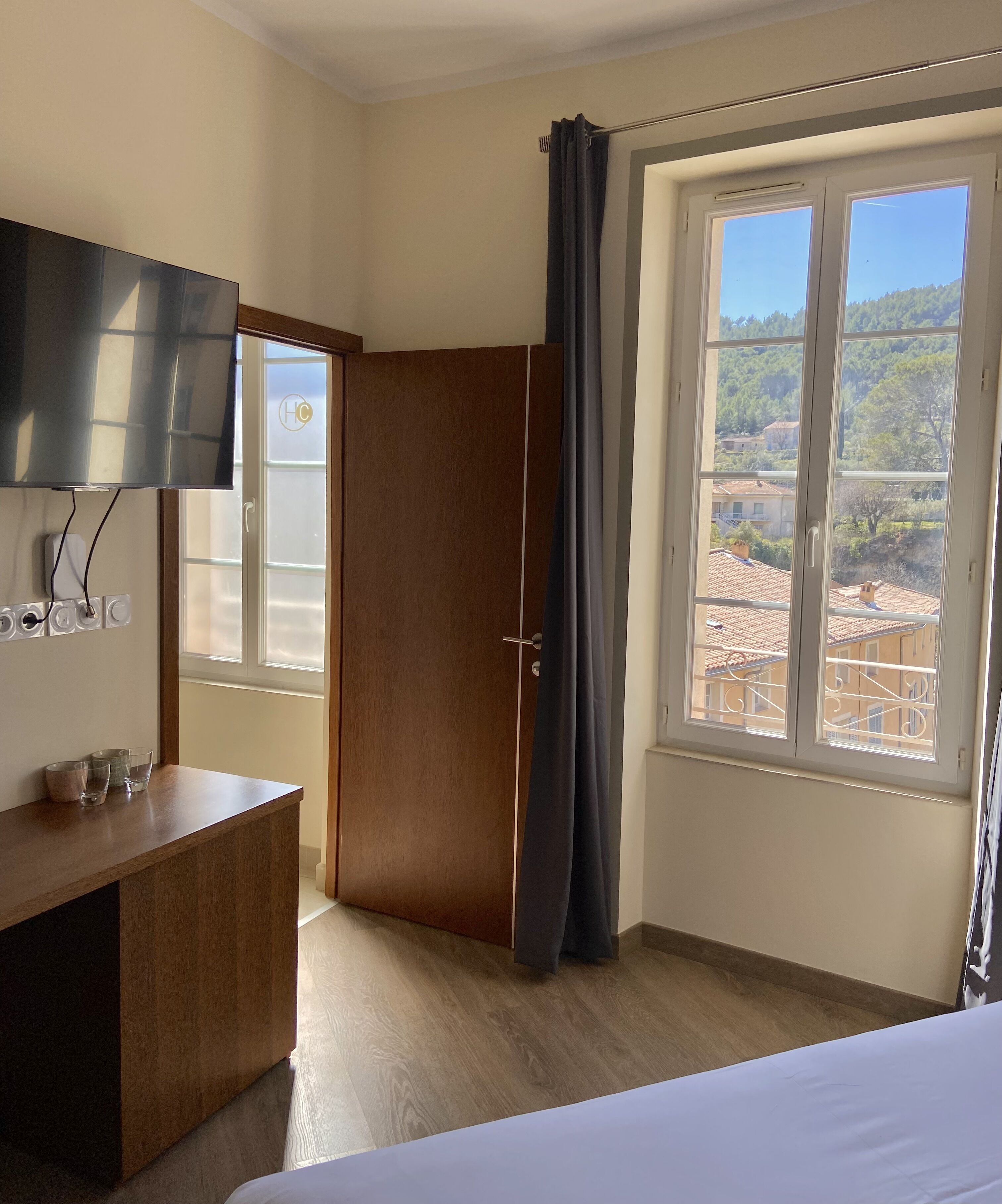 Une chambre à coucher comprenant un lit, une armoire en bois, un téléviseur fixé au mur et une fenêtre donnant sur des immeubles et des arbres.