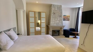 A bedroom with a large bed, a stone fireplace, a TV, and a view of the outside. - Hôtel La Falaise (Cotignac)