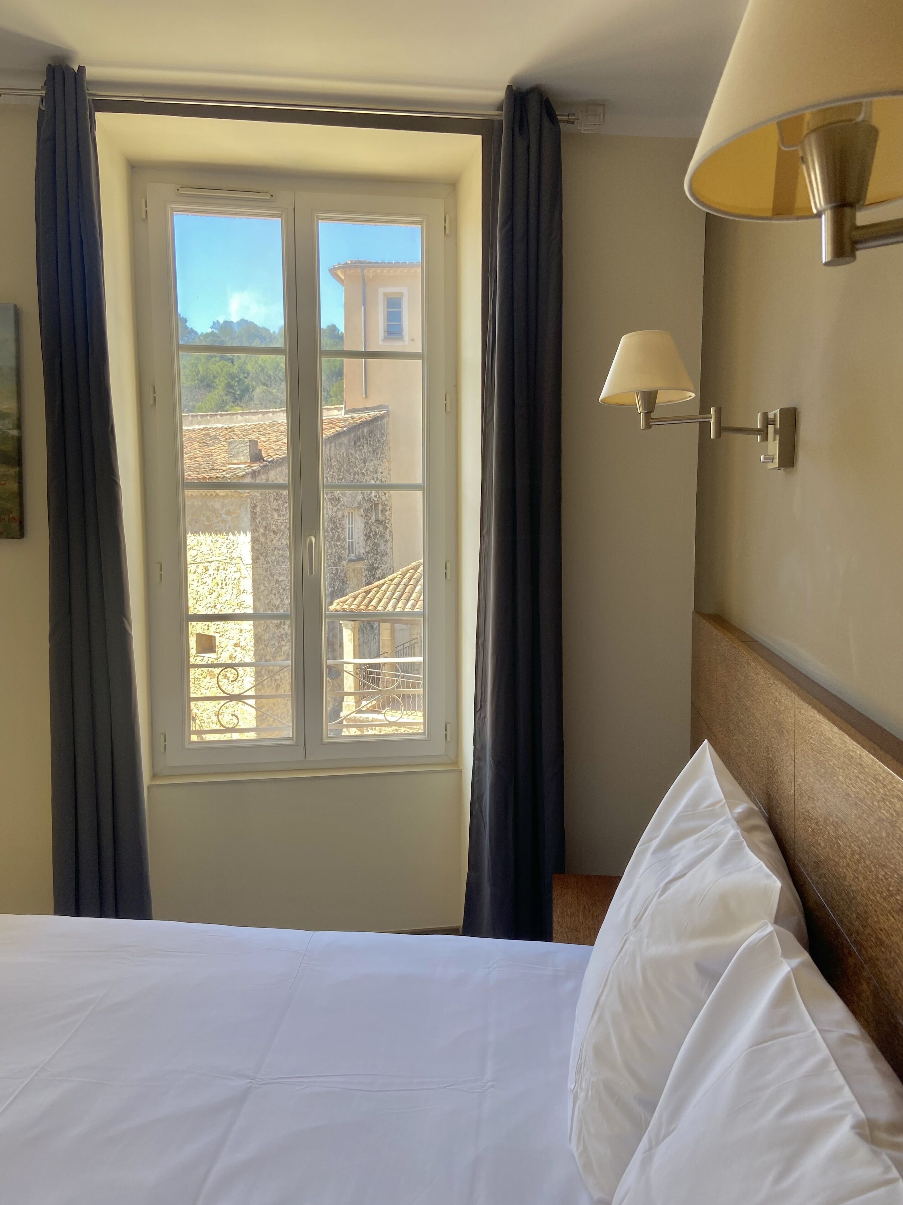 A hotel room with a bed, a window with curtains, and a wall-mounted lamp.