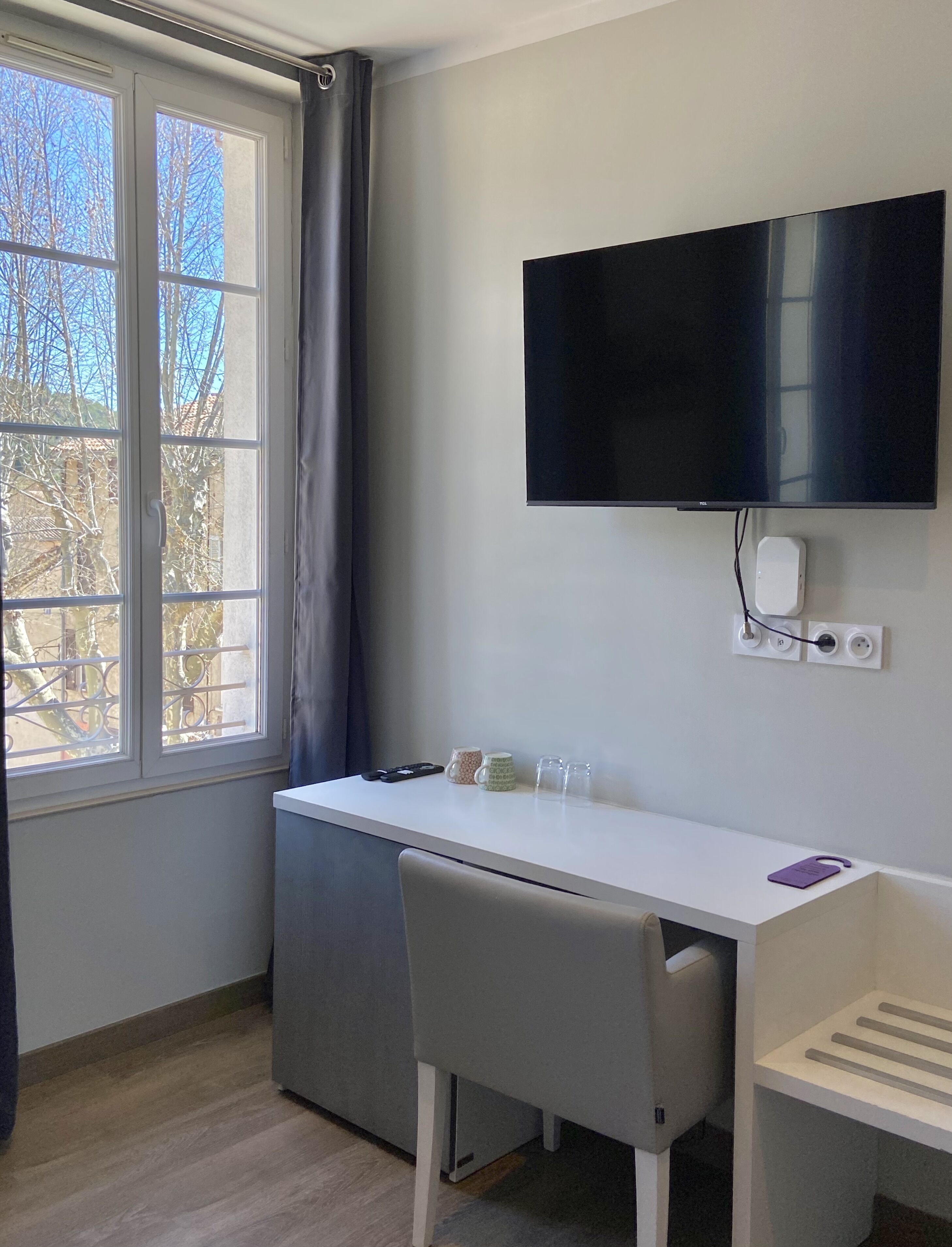 A small, modern room with a white desk, a grey chair, a flat-screen TV mounted on the wall, and a window with a view of trees and buildings.