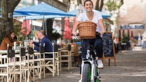 A person riding a bicycle with a basket, passing by outdoor seating areas and trees. - Hôtel La Falaise (Cotignac)